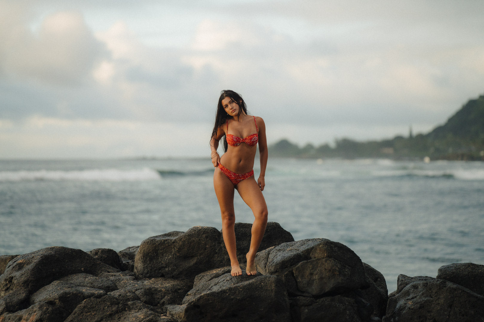 Woman in a red paisley bikini standing on rocks by the ocean with a cloudy sky.
